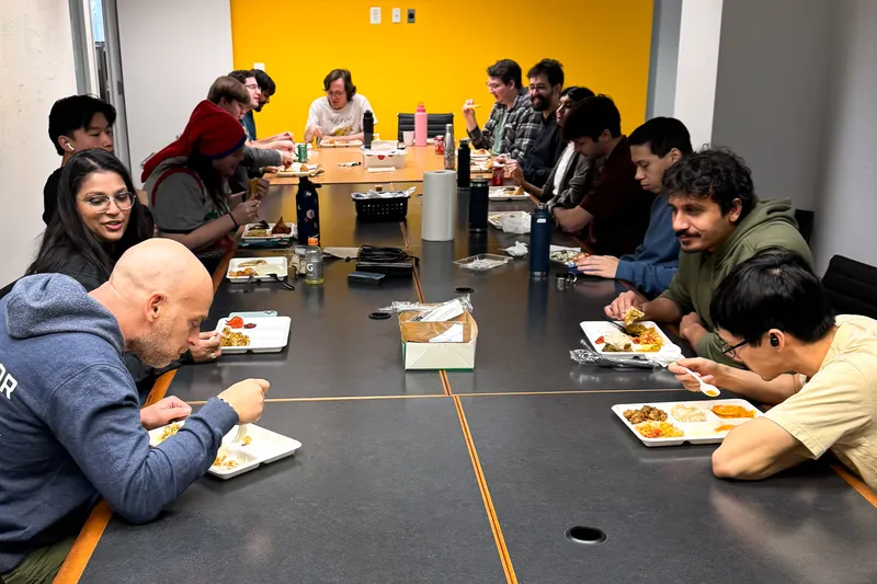 A large group of EAi teammates seated together at a long table, eating a catered lunch and talking.