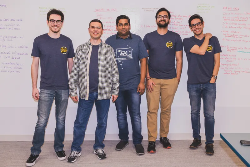 Five EAi teammates posing together in front of a whiteboard, most wearing matching event T-shirts.